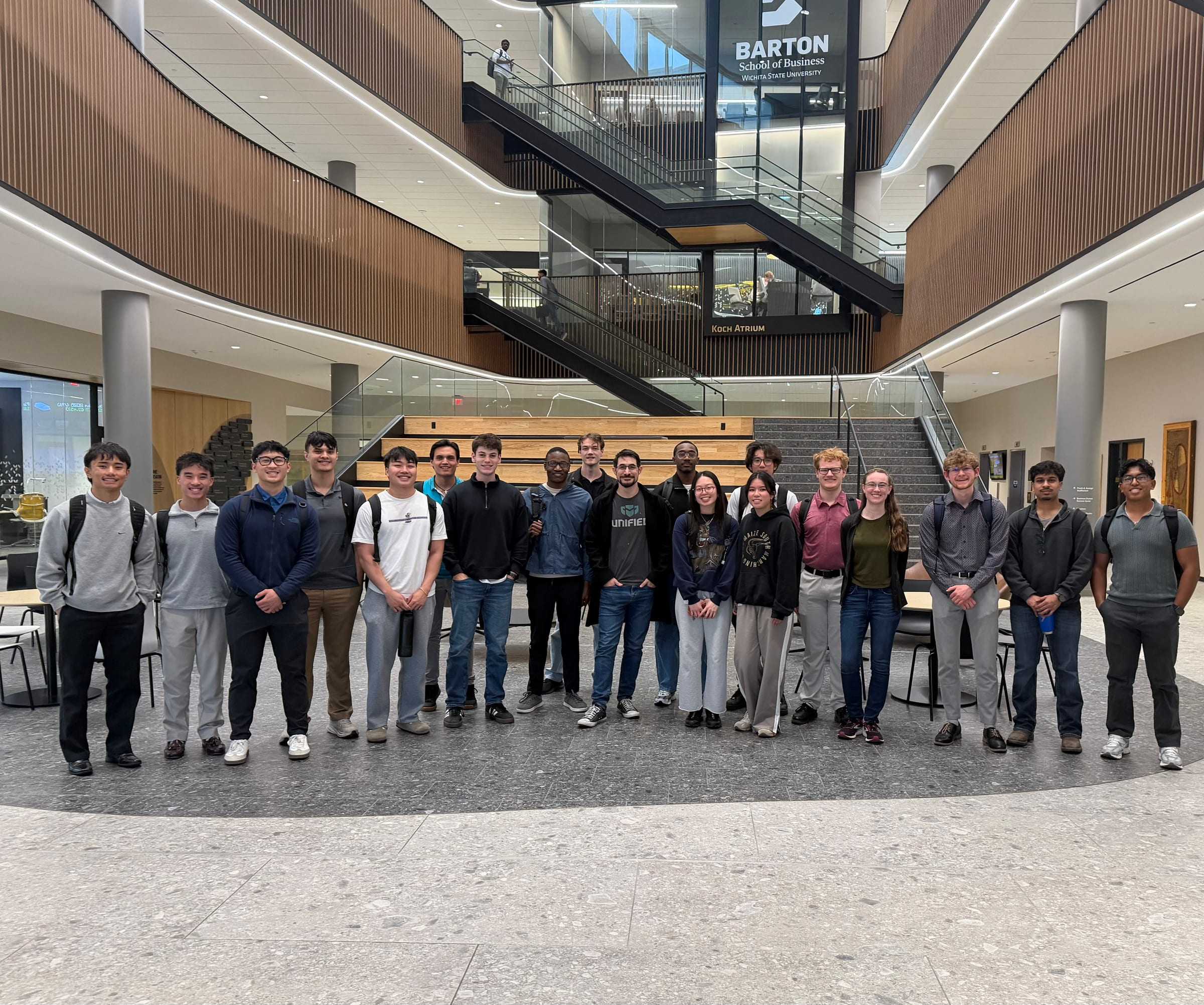 About 20 AI Club members stand in a row in the Koch Atrium of the Barton School of Business, under the Barton sign and the atrium stairs.