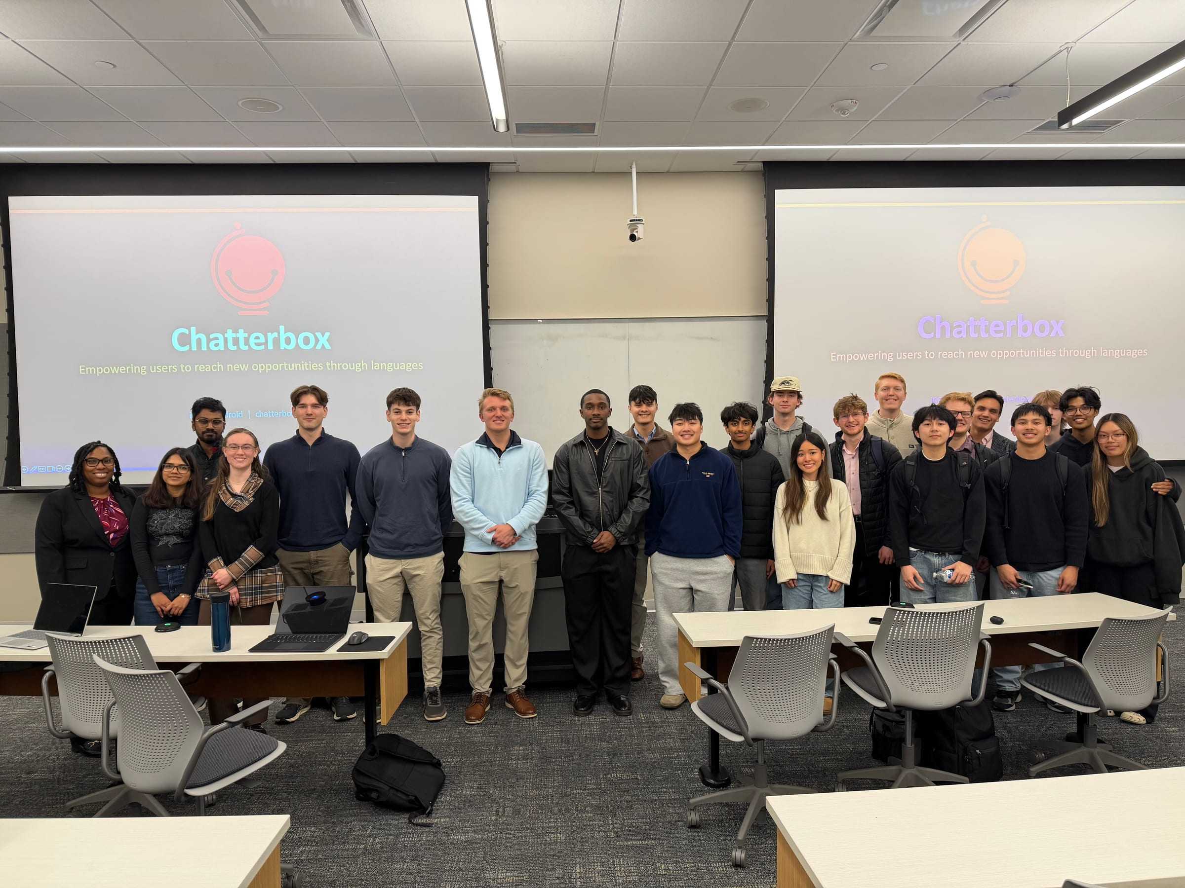 About two dozen people stand in front of two screens showing Chatterbox, a student-built language-learning app.