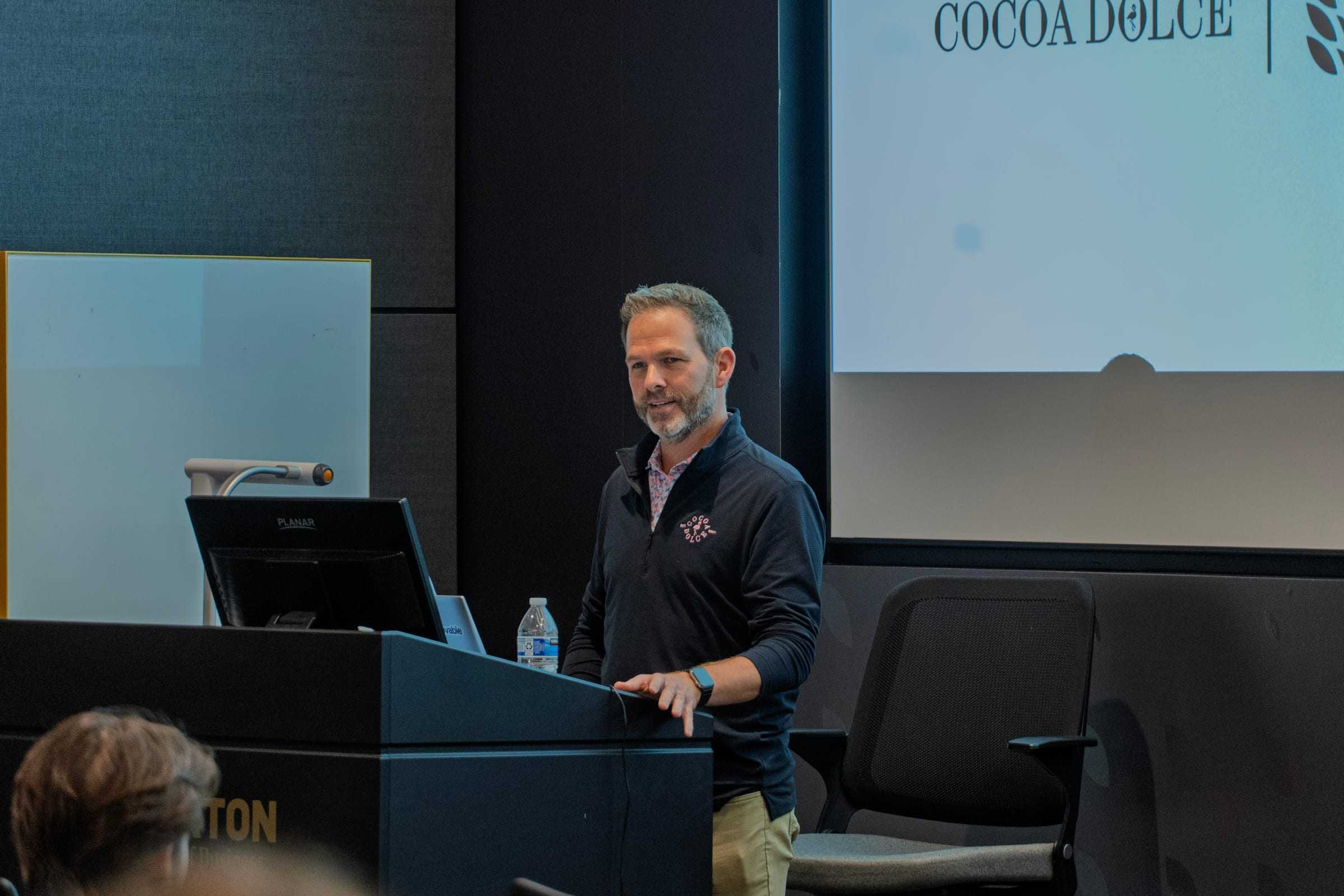 A speaker at the Woolsey Hall podium addresses the Chocolathon, with the Cocoa Dolce logo on the screen behind him.
