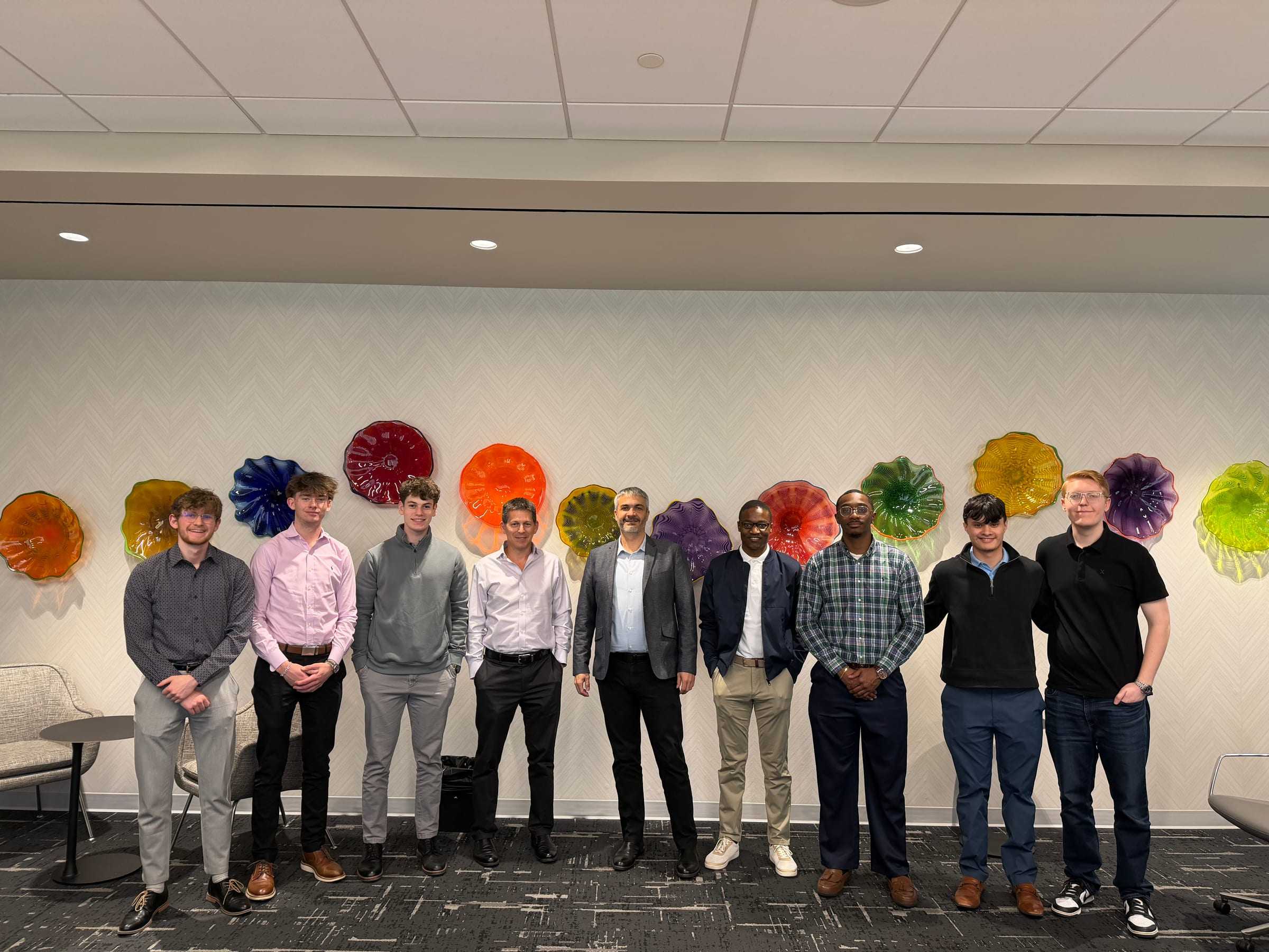 Nine people stand in front of a wall of colorful blown-glass art during an AI Club industry visit.